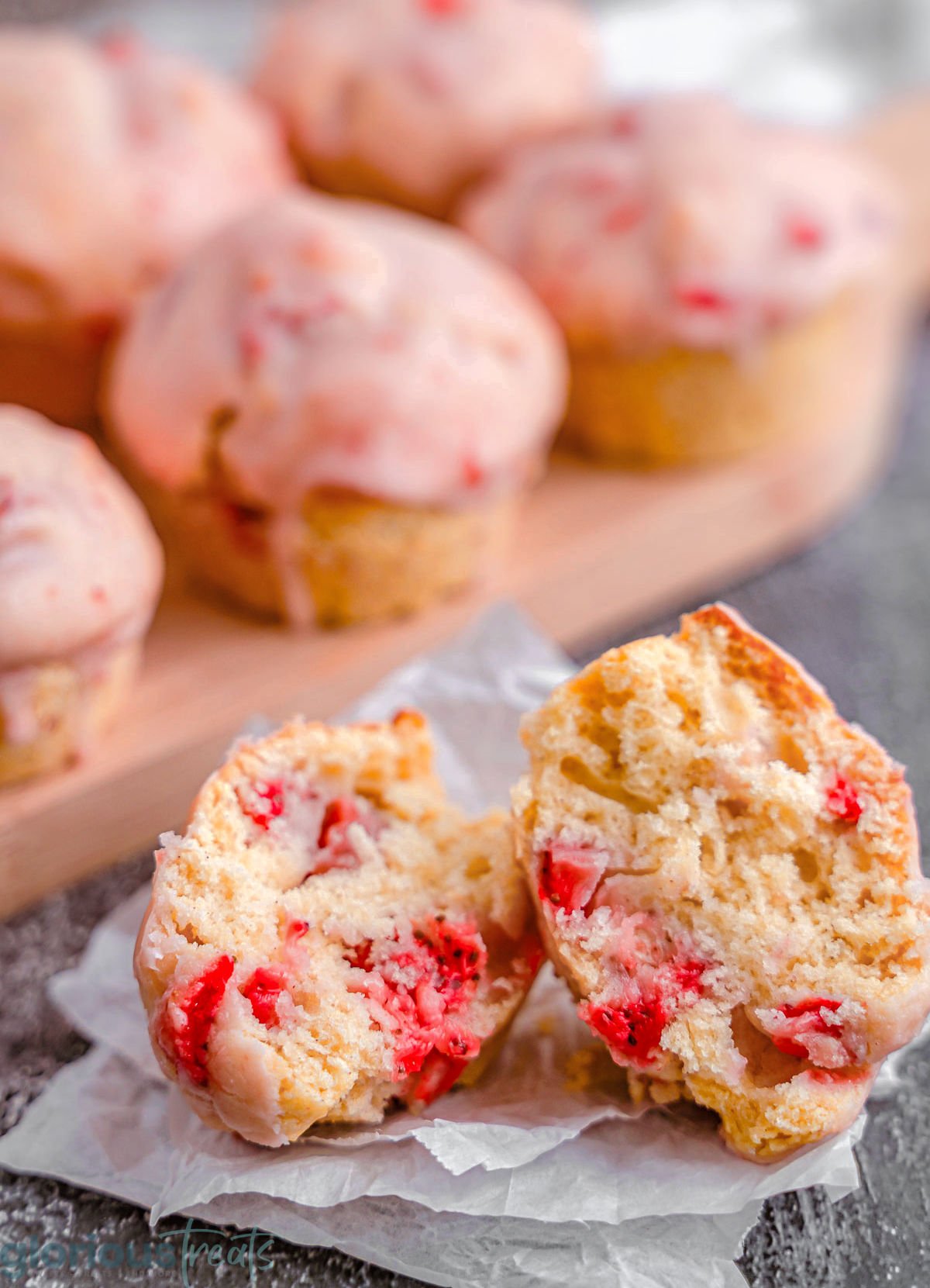 strawberry muffins with glaze on a cutting board. One muffin is broken in half to show the texture of the inside of the muffin.