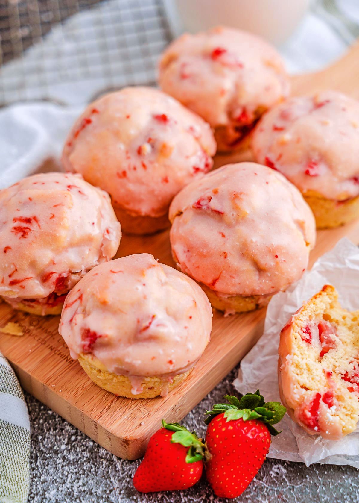 six strawberry muffins sitting on a wood board topped with a strawberry glaze. two fresh strawberries are sitting next to the muffins.