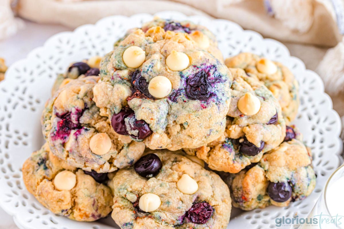 white plate with scalloped edges piled high with blueberry cookies that are studded with white chocolate chips. fresh blueberries are scattered about the plate.