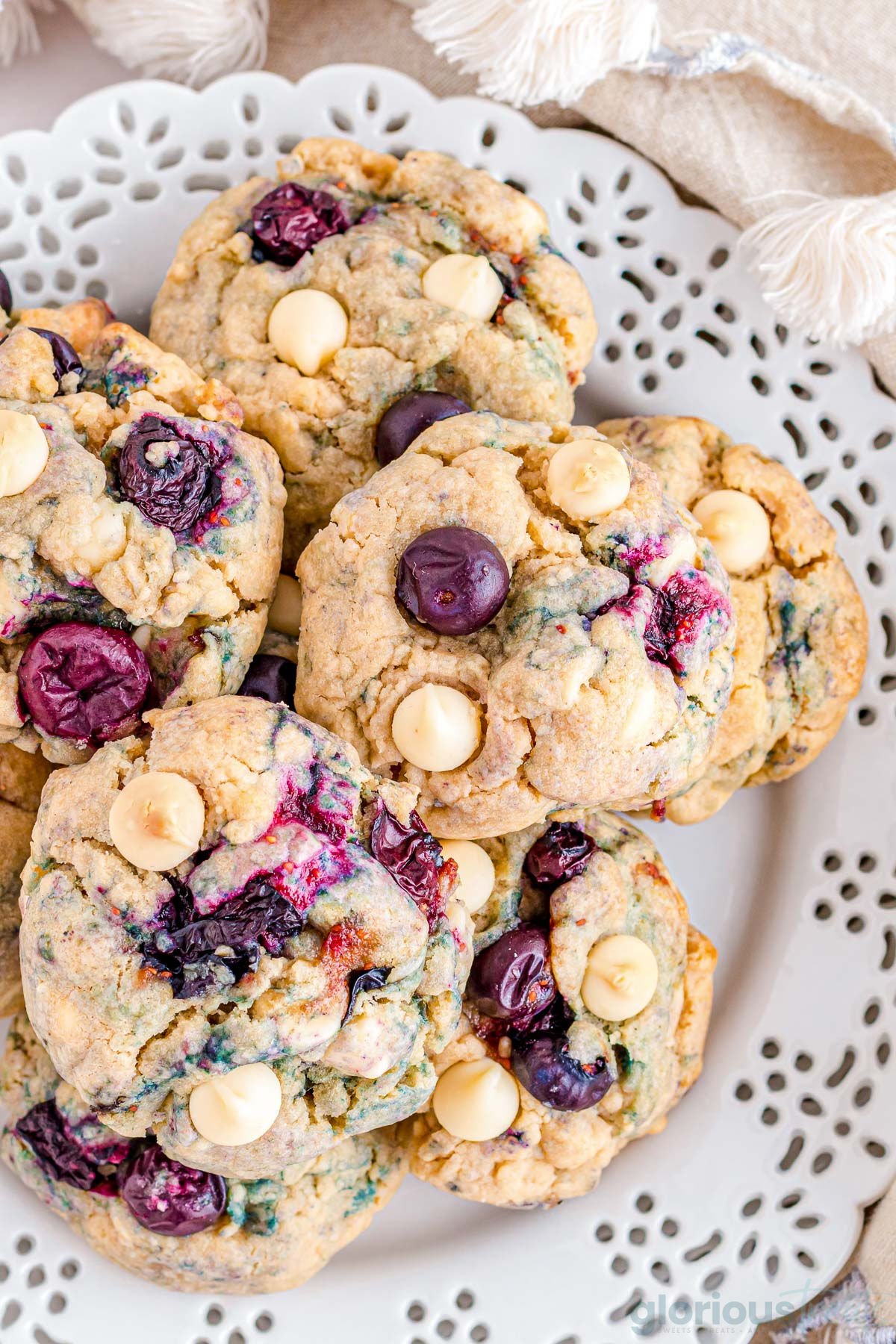 blueberry cookies are arranged on a white plate. cookies are close up to show the texture of the cookies and detail of blueberry and white chocolate chips.