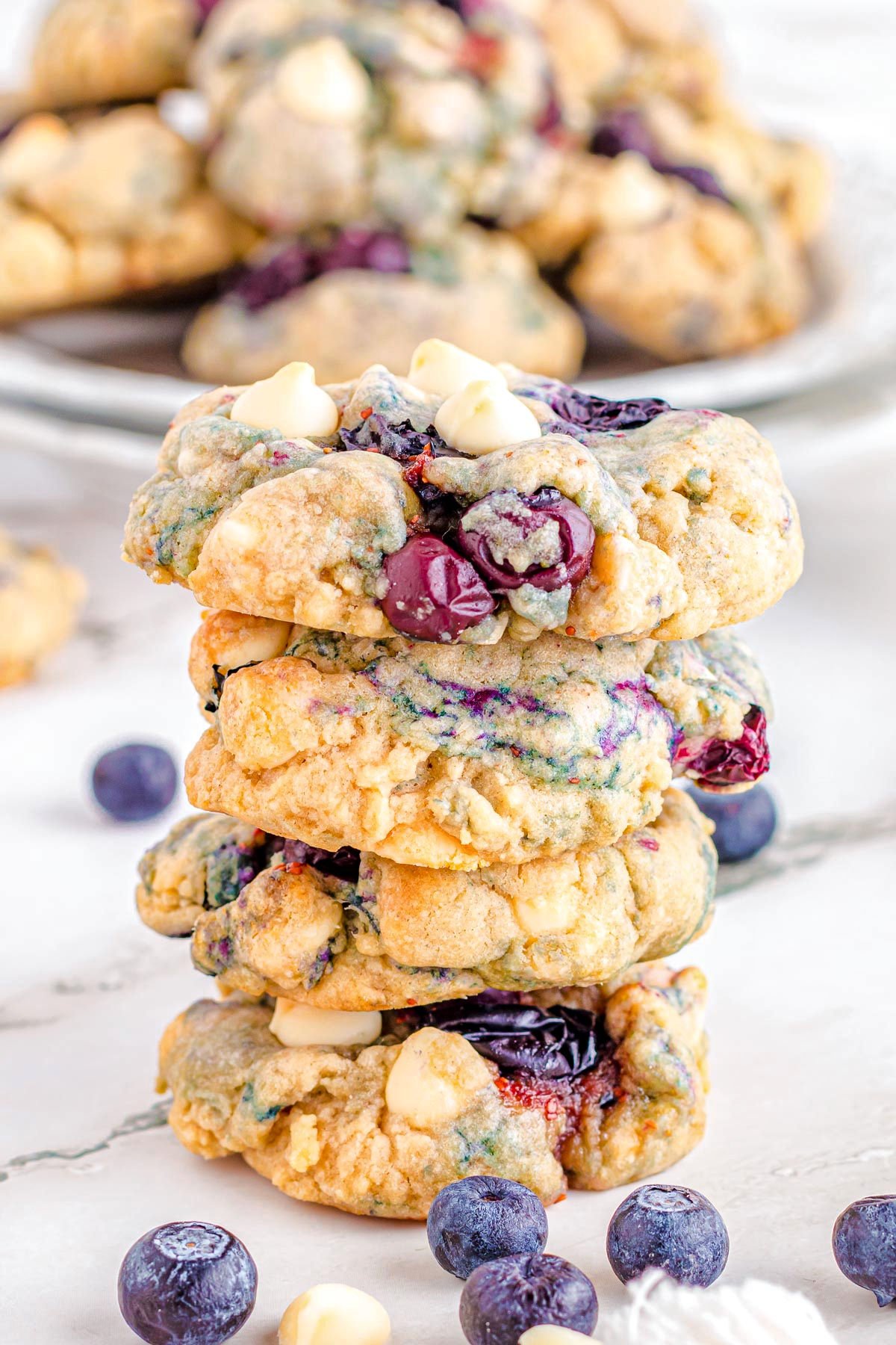 four blueberry cookies are stacked on a table. bluberries and white chocolate chips are scattered on the table in front of the cookies.