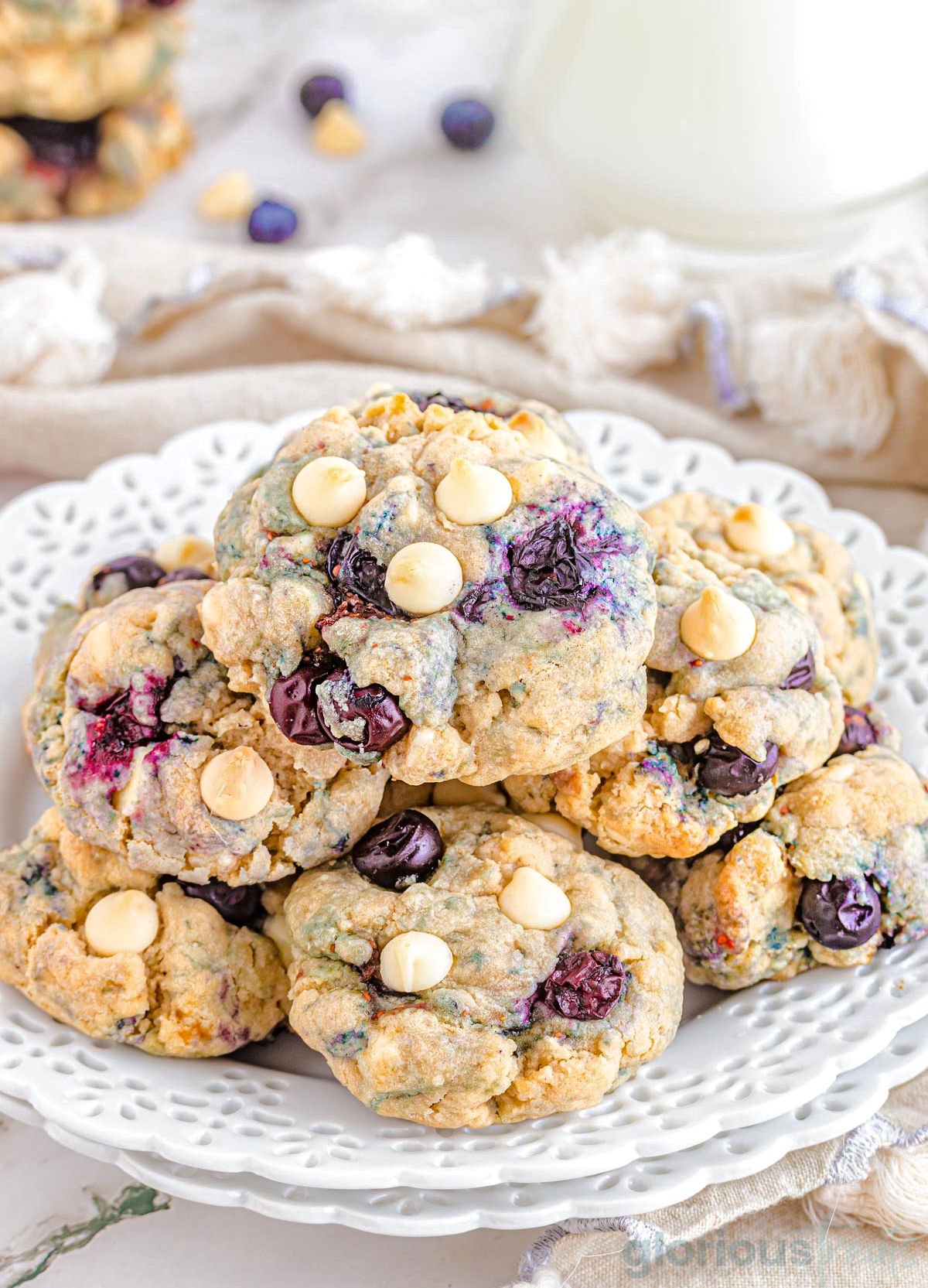white plate with scalloped edges piled high with blueberry cookies that are studded with white chocolate chips. fresh blueberries are scattered about the plate. 