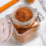 top down view of small spice jar filled with homemade pumpkin pie spice with a hinged lid. cinnamon stick and whole nutmeg can be seen in background. jar is sitting on marble surface.