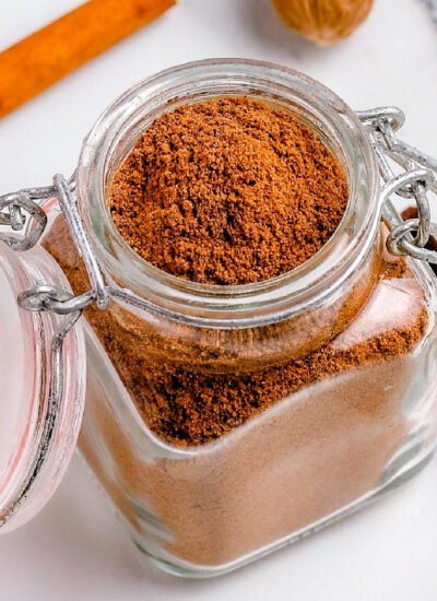 top down view of small spice jar filled with homemade pumpkin pie spice with a hinged lid. cinnamon stick and whole nutmeg can be seen in background. jar is sitting on marble surface.