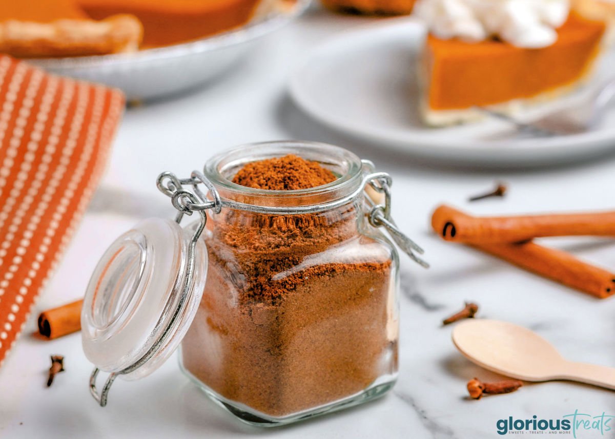 small spice jar filled with homemade pumpkin pie spice. slice of pumpkin pie on white round plate can be seen in background. cinnamon sticks to the right of the jar.