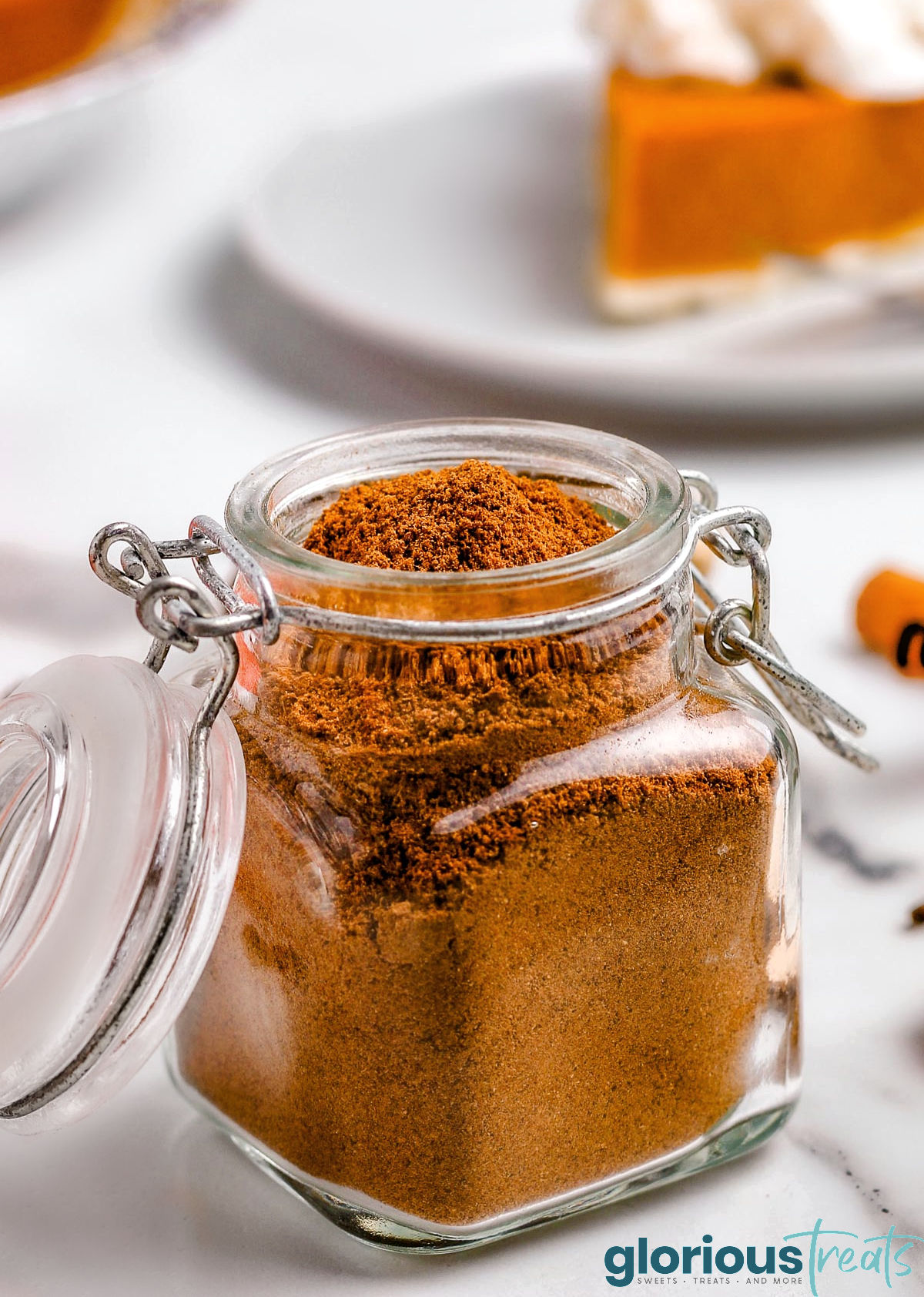 homemade pumpkin pie spice in small glass jar with hinged jar. whole nutmeg and cloves can be seen in the background.