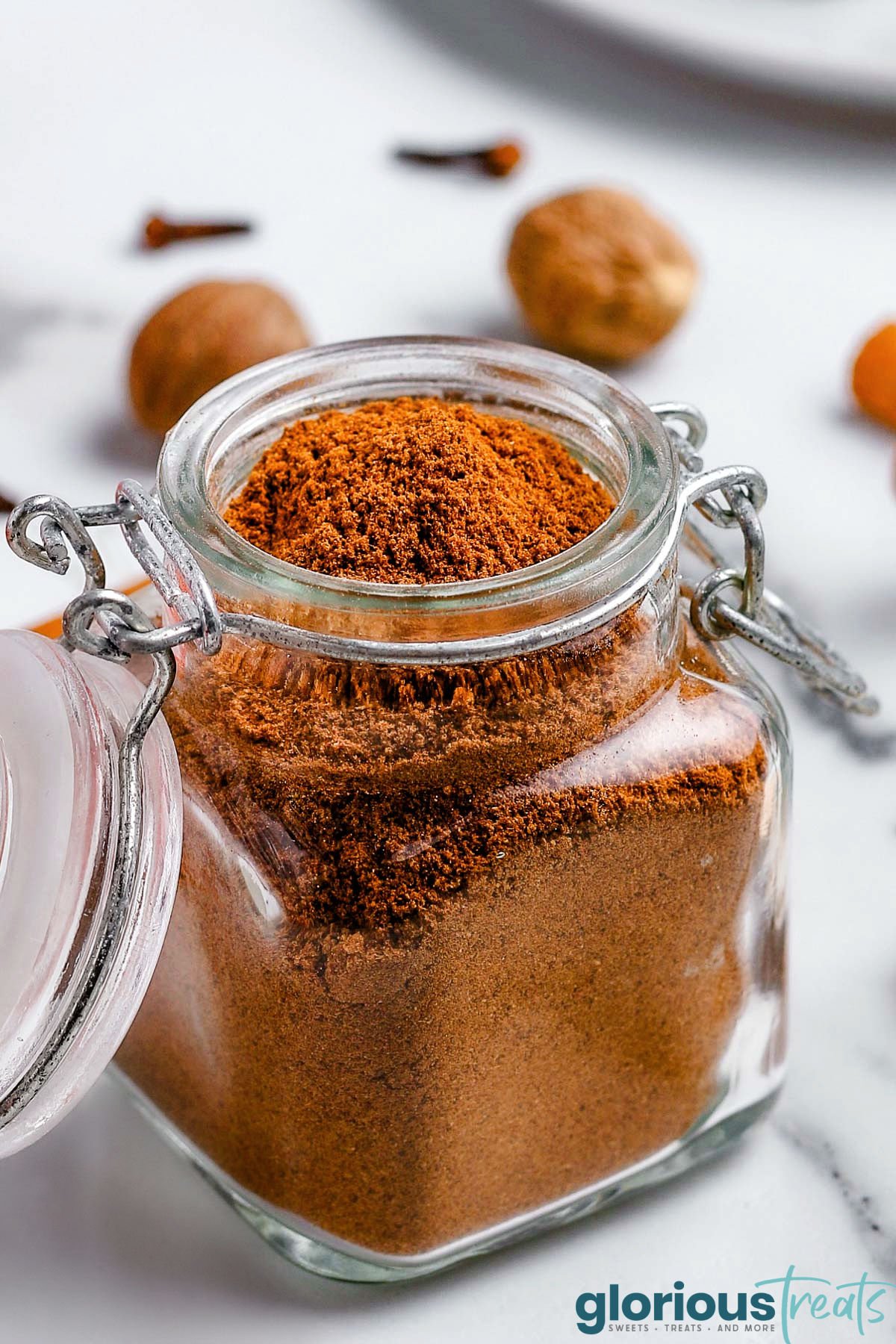 homemade pumpkin pie spice in small glass jar with hinged jar. whole nutmeg and cloves can be seen in the background.