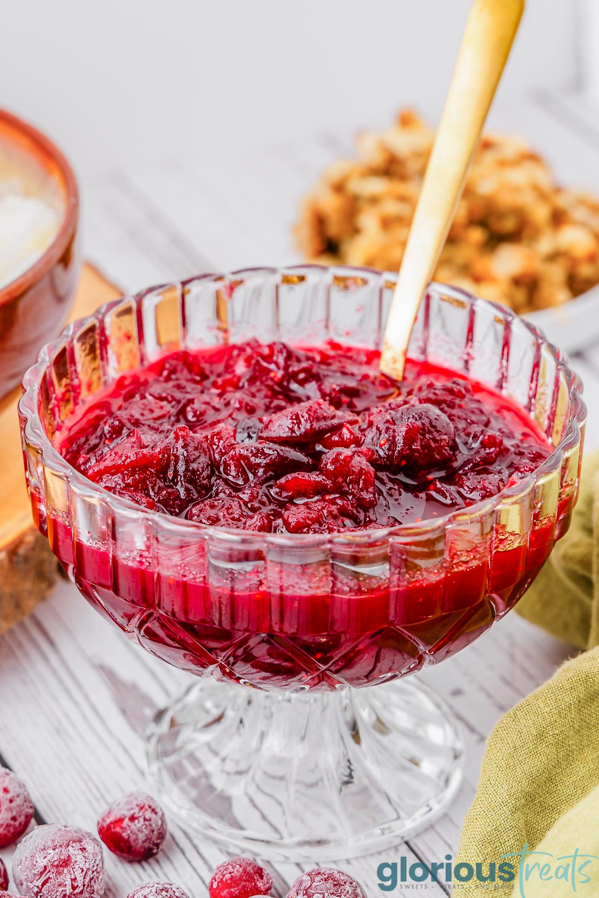 footed glass bowl filled with cranberry sauce with a gold spoon in the bowl.