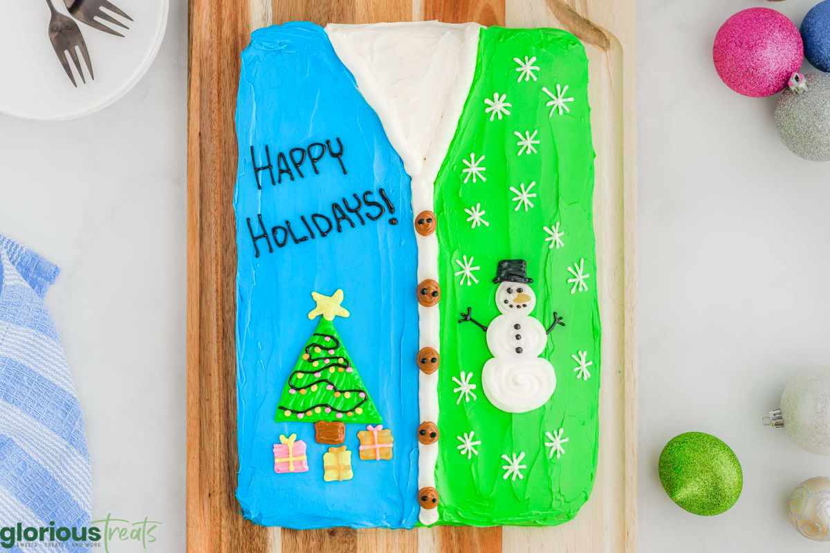 top down view of red velvet cake decorated to look like an ugly christmas sweater complete with snowman and christmas tree. cake is sitting on wood cutting board.