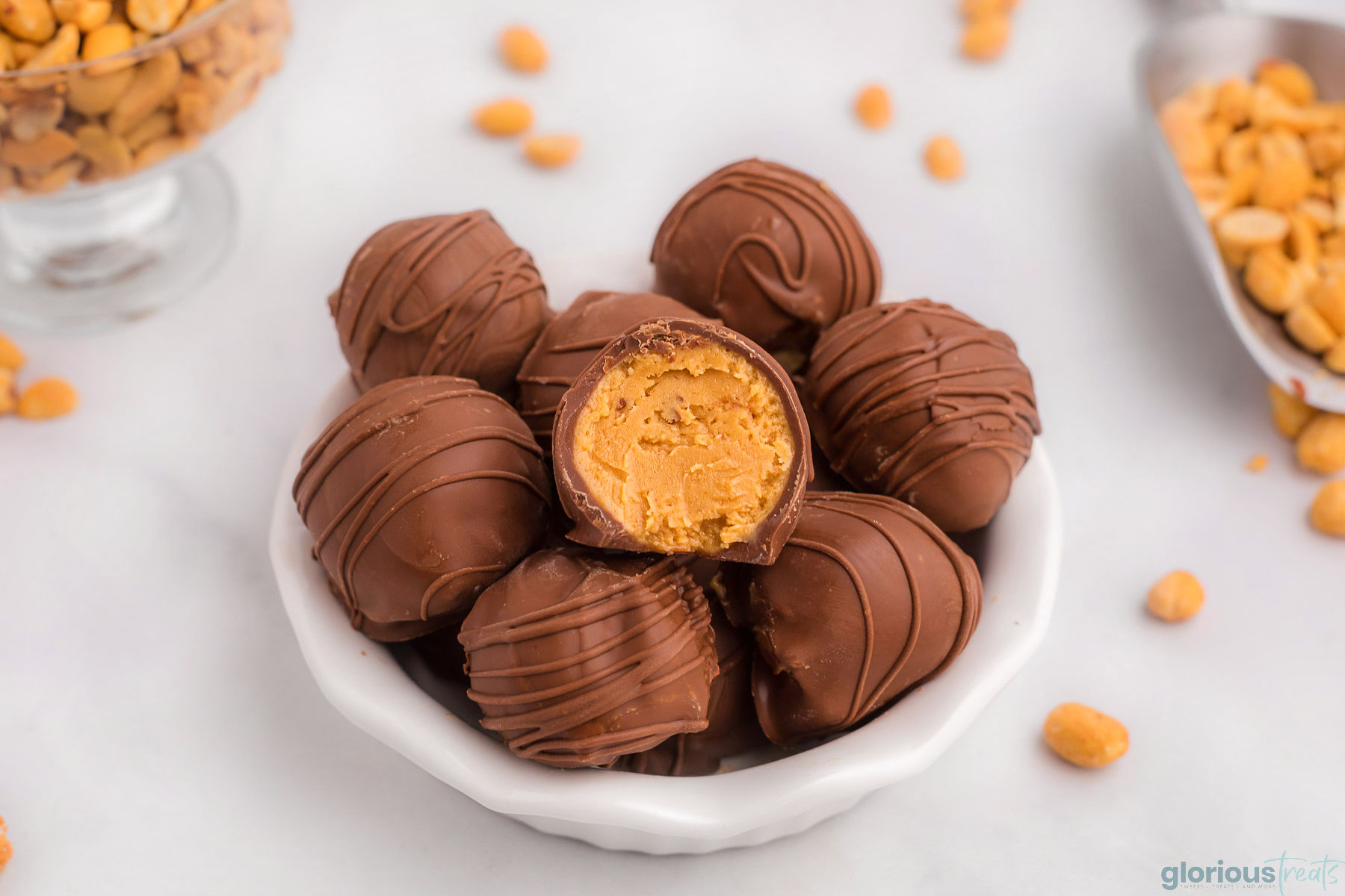 wide shot of a small, white bowl with scalloped edges loaded to overflowing with homemade peanut butter balls recipe. top ball has a bite taken out of it. peanuts are scattered around the bowl on the white surface it is sitting on.