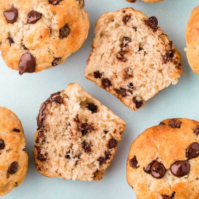 top down view of banana chocolate chip muffins with one muffin split in half showing the chocolate chips on the inside. light aqua background.