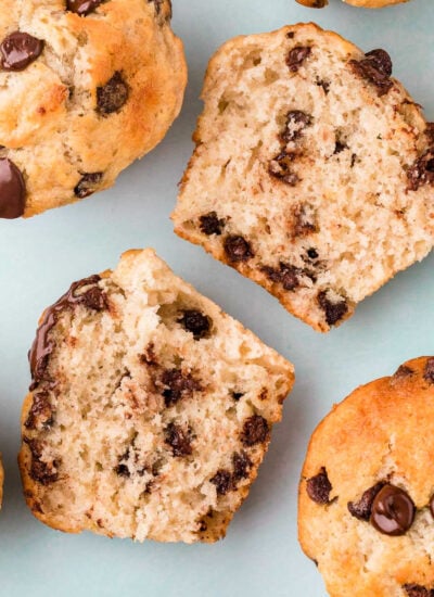 top down view of banana chocolate chip muffins with one muffin split in half showing the chocolate chips on the inside. light aqua background.