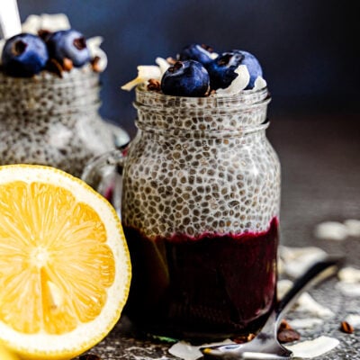 chia pudding in a mason jar glass with half a lemon sitting next to it. the pudding is topped with fresh blueberries and coconut flakes and the pudding is layered with blueberry compote.