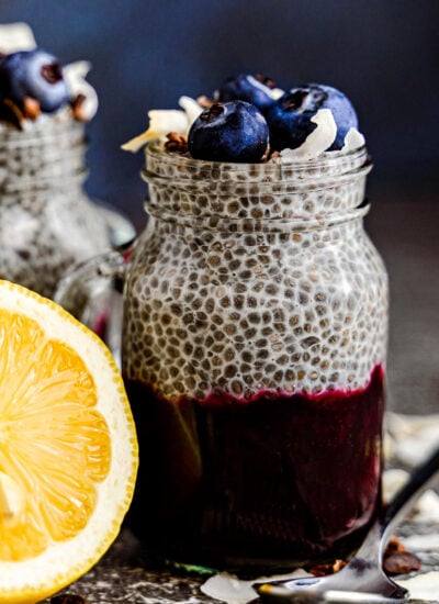 chia pudding in a mason jar glass with half a lemon sitting next to it. the pudding is topped with fresh blueberries and coconut flakes and the pudding is layered with blueberry compote.