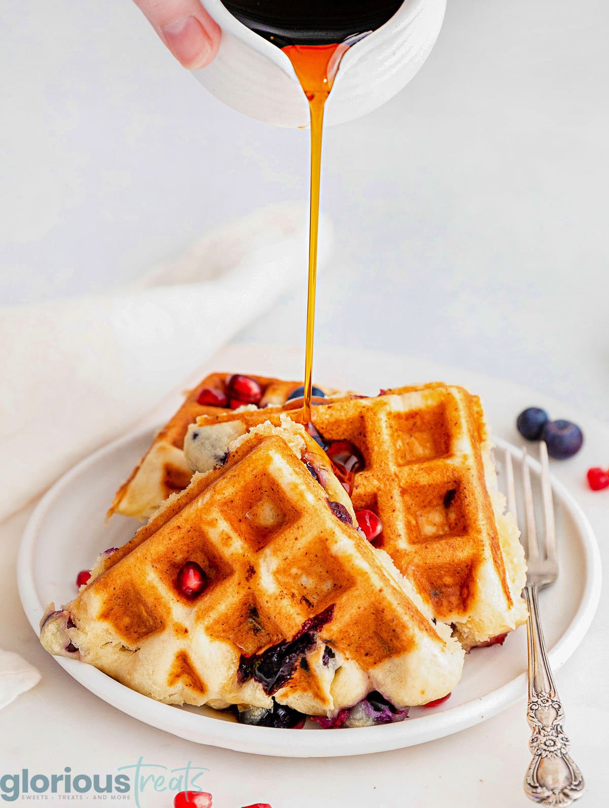 Syrup being drizzled over three blueberry waffles on a round white plate with a fork next to the waffles.