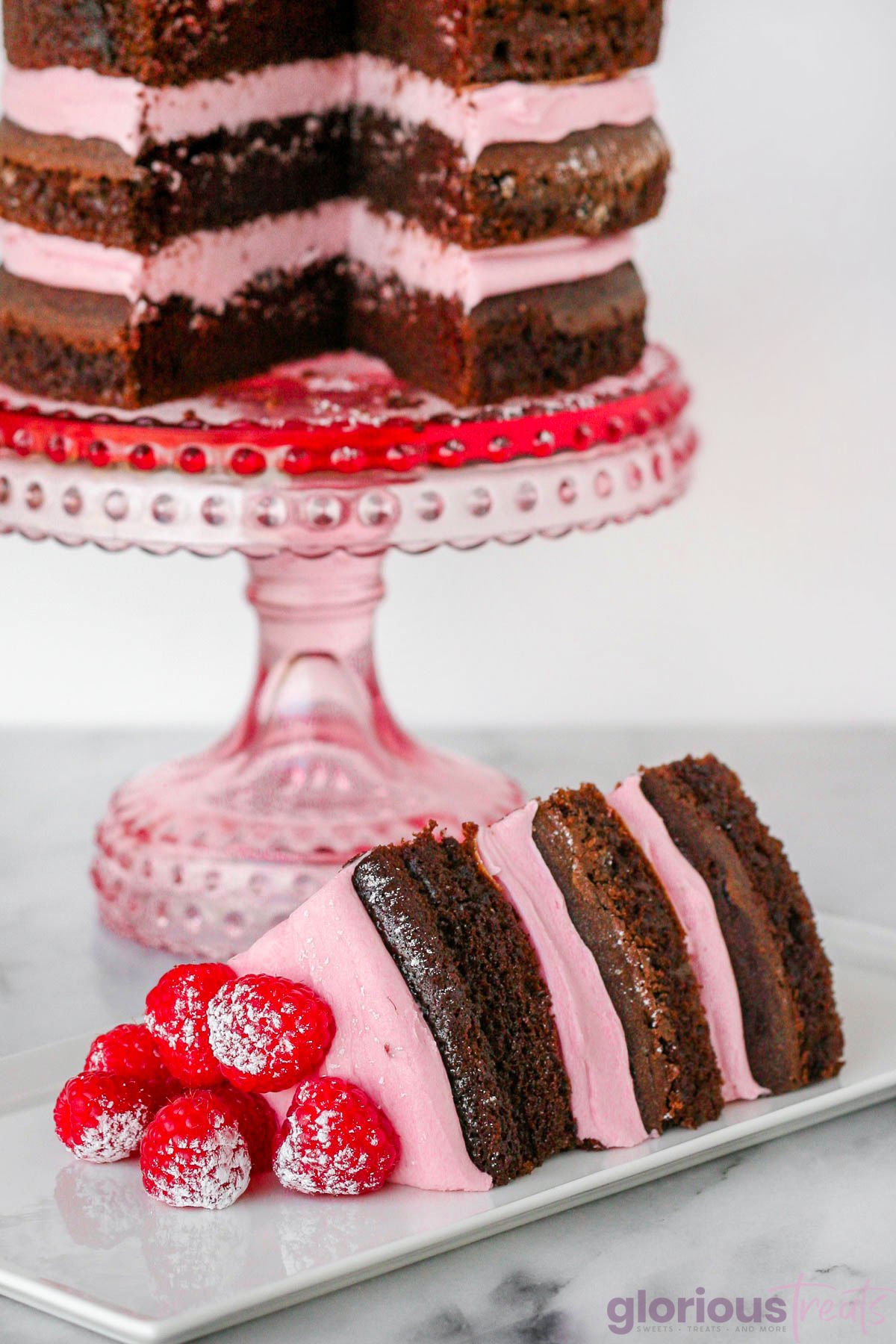 slice of chocolate cake with raspberry cream cheese frosting on plate and the rest of the cake can be seen on a pink cake stand in the background. The cake has been garnished with raspberries and powdered sugar.
