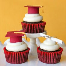 three chocolate cupcakes with buttercream frosting on individual cupcake pedestals topped with fondant graduation caps and tassels.