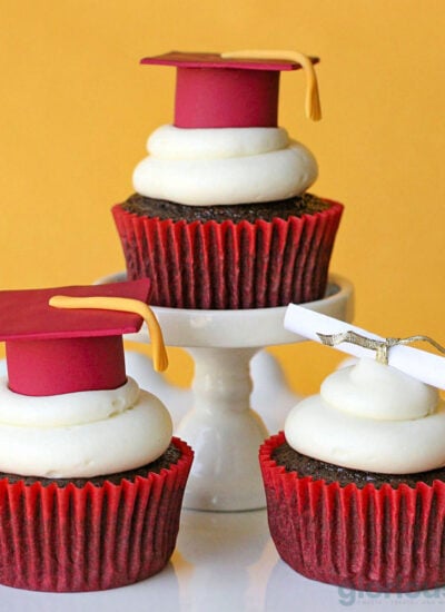 three chocolate cupcakes with buttercream frosting on individual cupcake pedestals topped with fondant graduation caps and tassels.