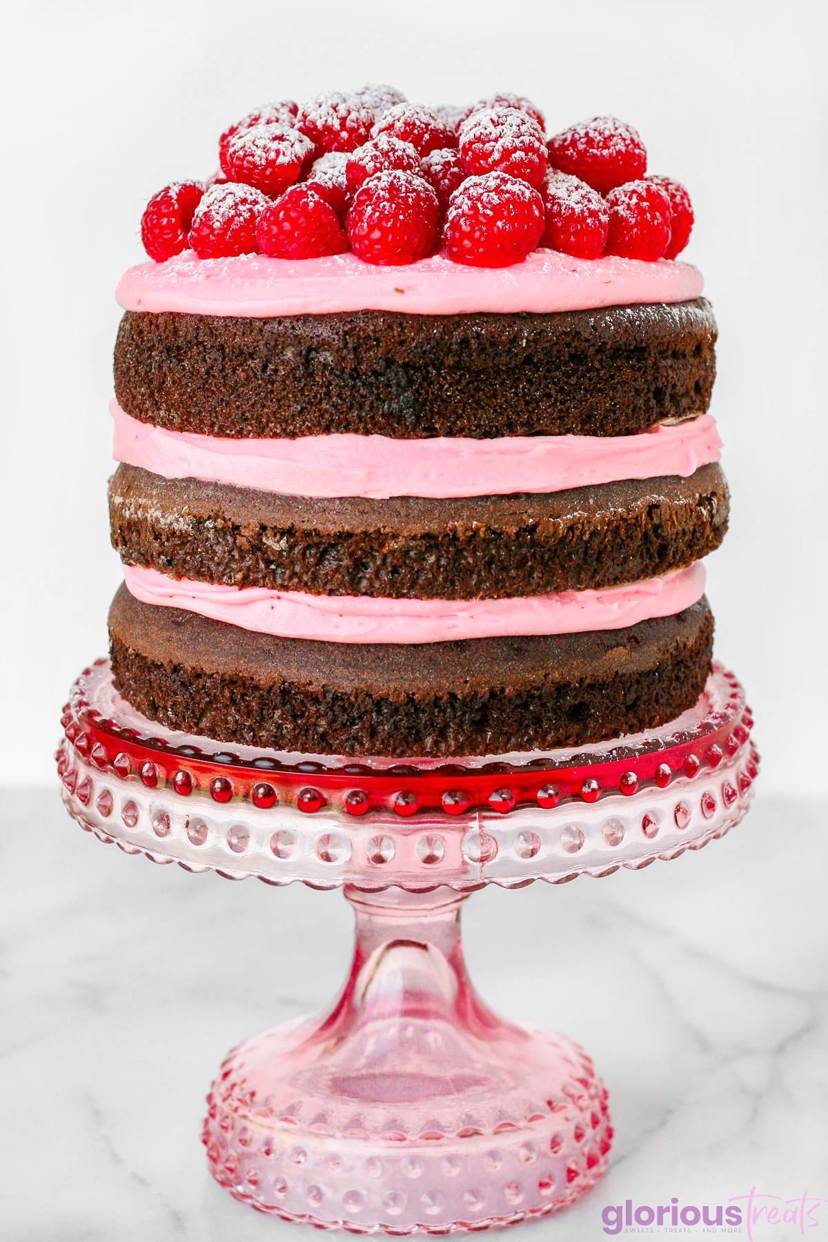 three layer raspberry chocolate cake with raspberry buttercream between each cake layer. cake is on pink cake stand and topped with fresh raspberries and powdered sugar.