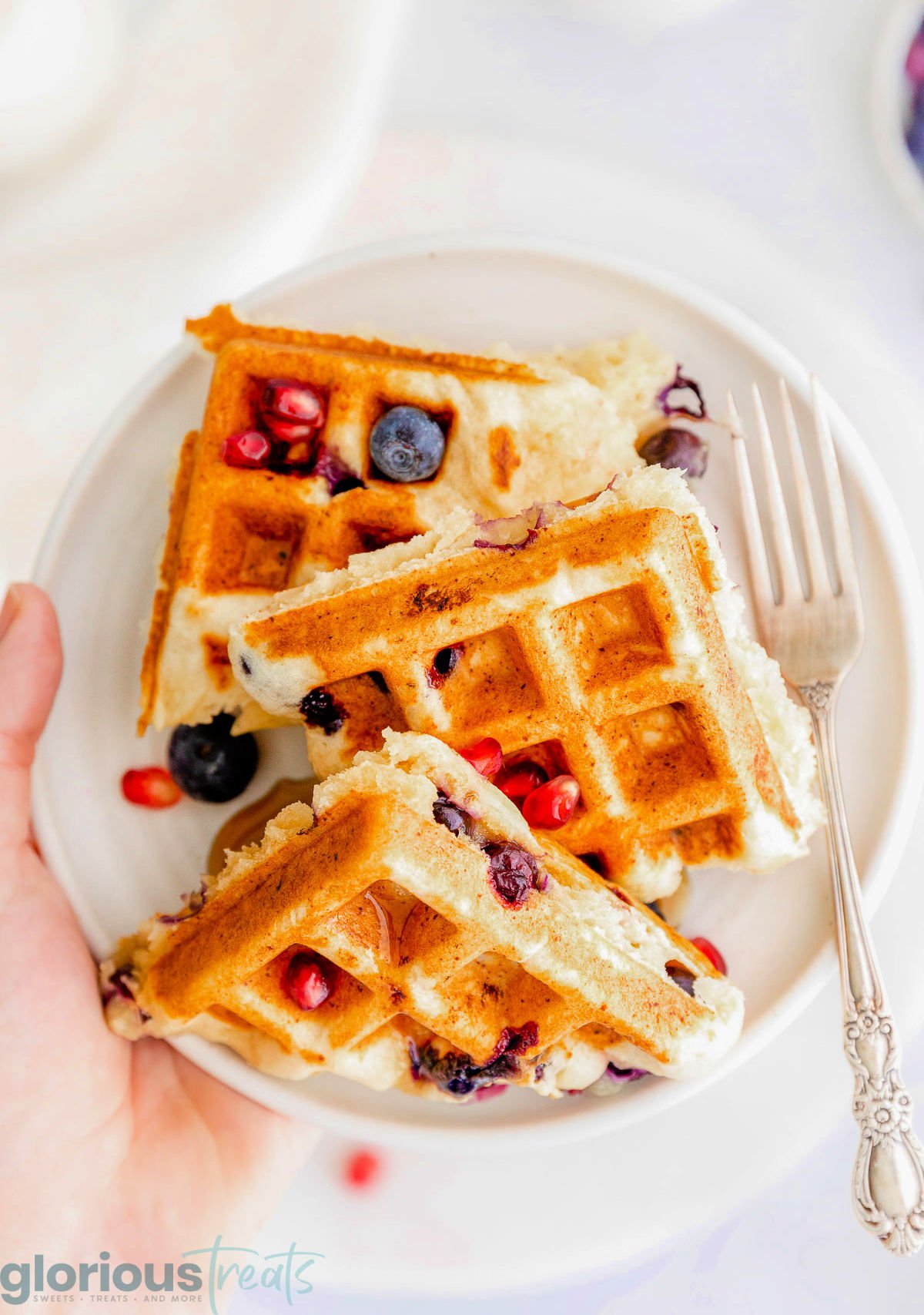 white plate with three blueberry ricotta waffles on it being held up over a white surface. 