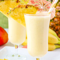 two light yellow fruit smoothies garnished with pineapple slices, paper umbrellas, and paper straws. Fresh pineapple slices, bananas and mangoes can be seen in the background.