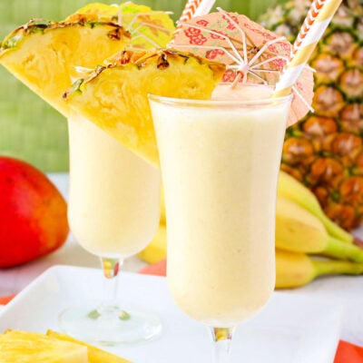 two light yellow fruit smoothies garnished with pineapple slices, paper umbrellas, and paper straws.