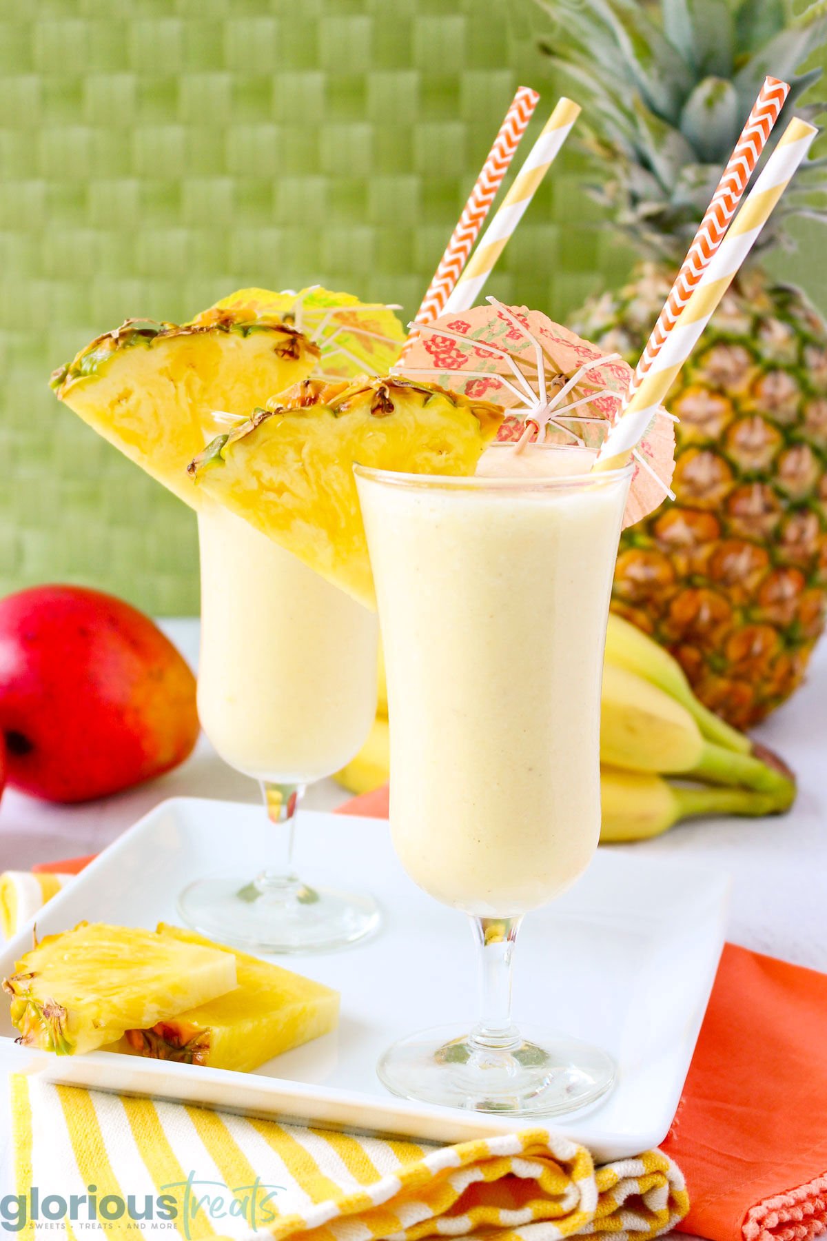 two fruit smoothies on a white serving platter with pineapple slices and bananas, pineapples, and more fruit in the background.