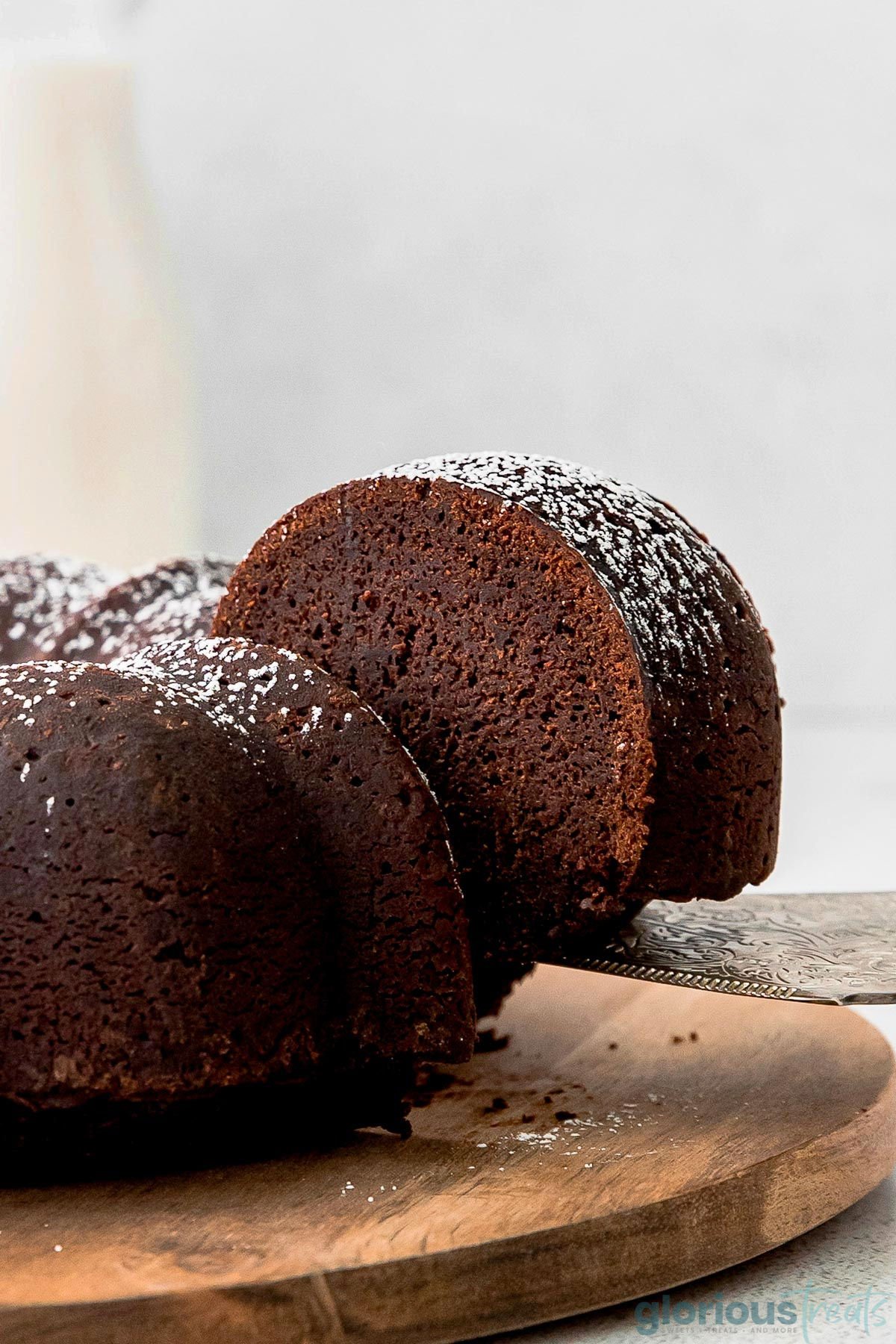 a chocolate bundt cake on a wood cutting board with a spatula removing a slice of cake from it.