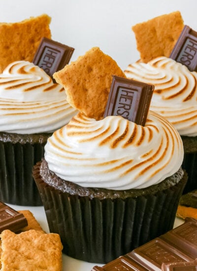 three chocolate s'mores cupcakes topped with a hershey square and graham crackers and extra toppings on the white surface beneath them.