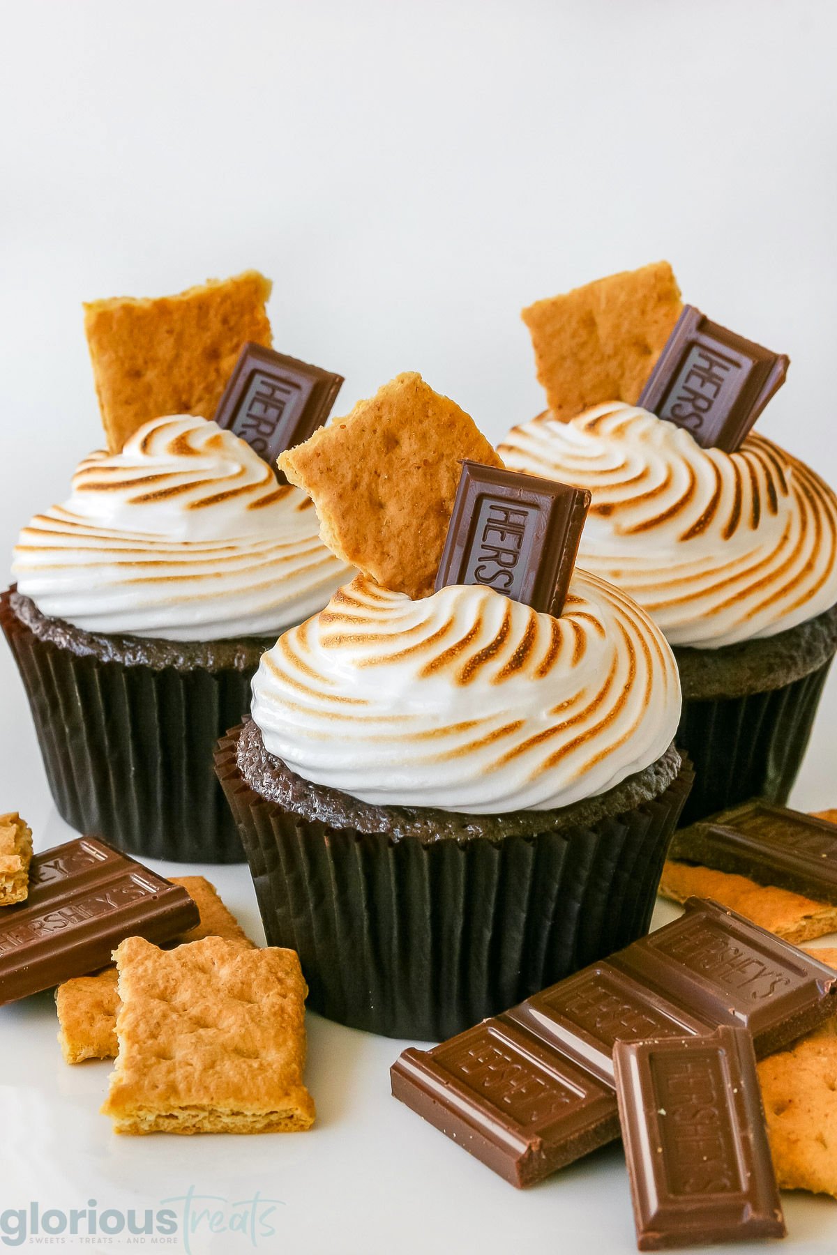three chocolate s'mores cupcakes topped with a hershey square and graham crackers and extra toppings on the white surface beneath them.