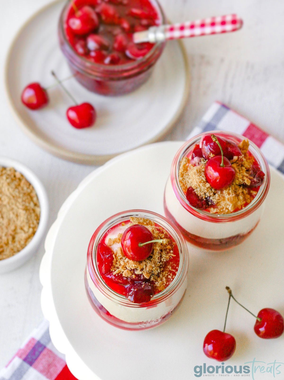 an overhead photo of two cherry cheesecake portions on a white circular serving platter and a jar of cherries with a spoon in the jar in the upper left corner out of focus
