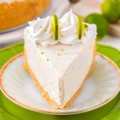Slice of key lime cheesecake on a white round plate sitting on a larger green plate. Cheesecake slice is topped with whipped cream and lime slices.