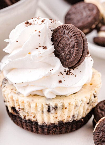 mini oreo cheesecake topped with whipped cream and a mini oreo sitting on a white surface with oreo cookies scattered about.