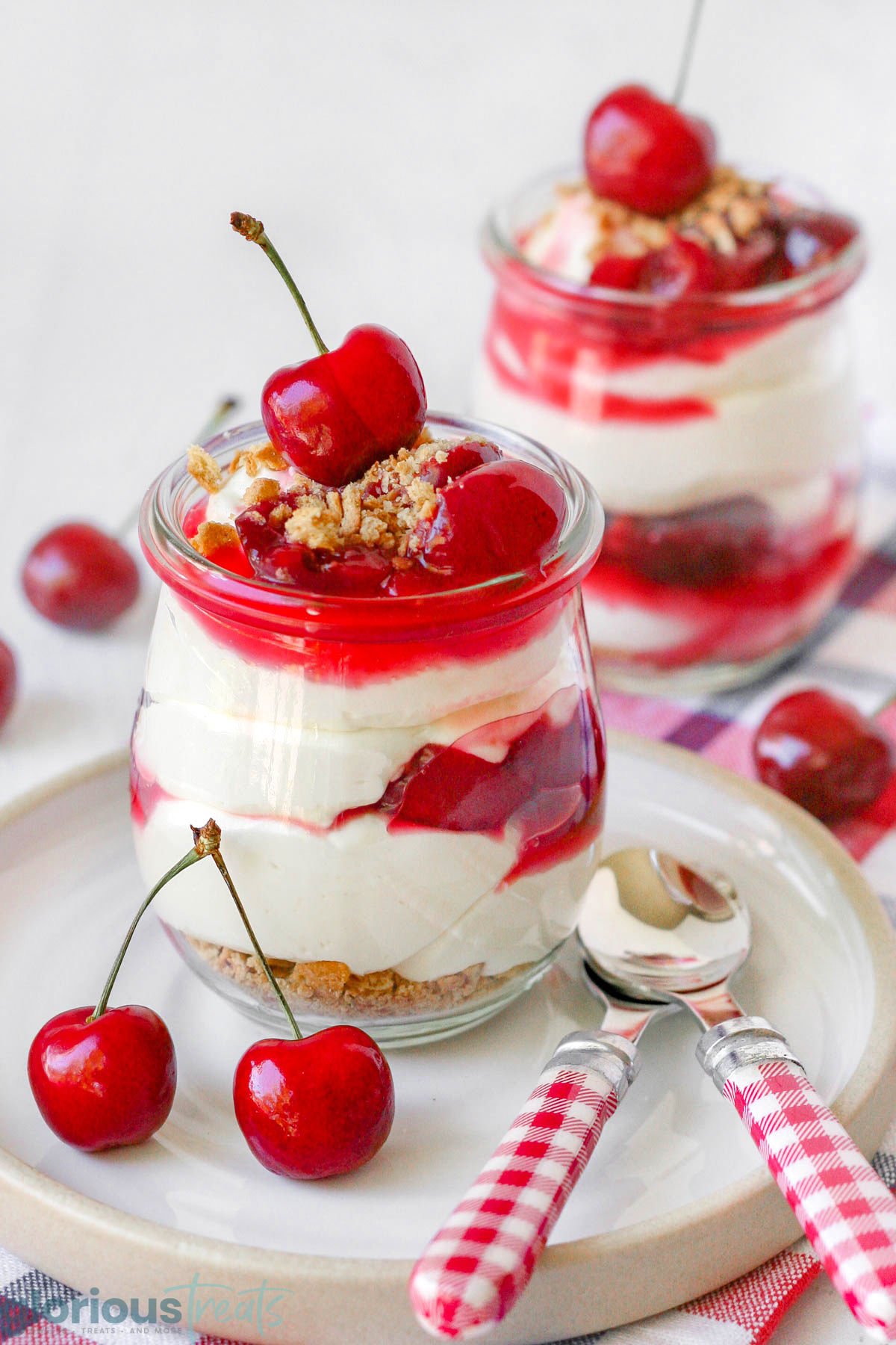 a white plate with a jar of cherry cheesecake on it along with two spoons and some cherries, and another jar out of focus in the background