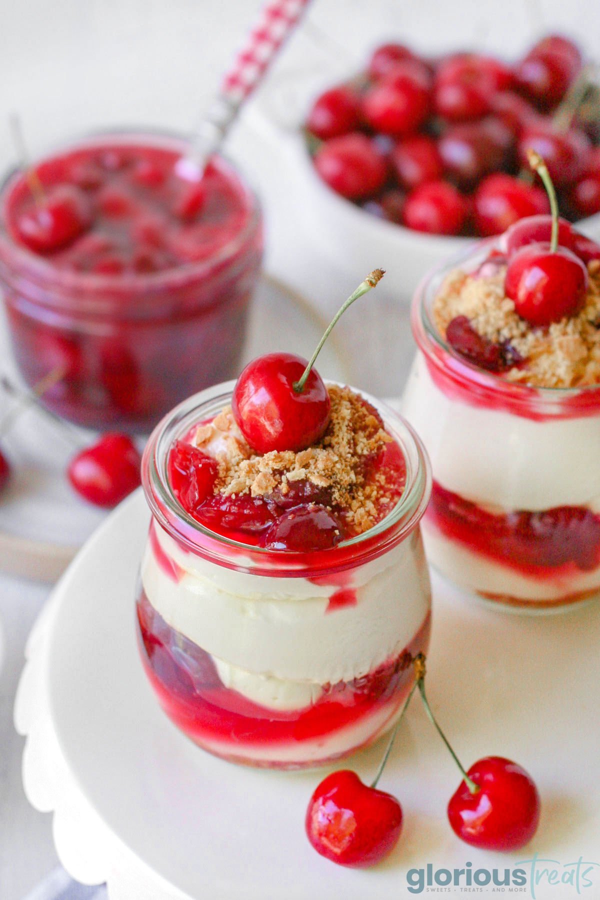 a couple jars of no bake cherry cheesecake topped with cherries, and a jar full of cherry topping in the background beside a bowl of whole cherries