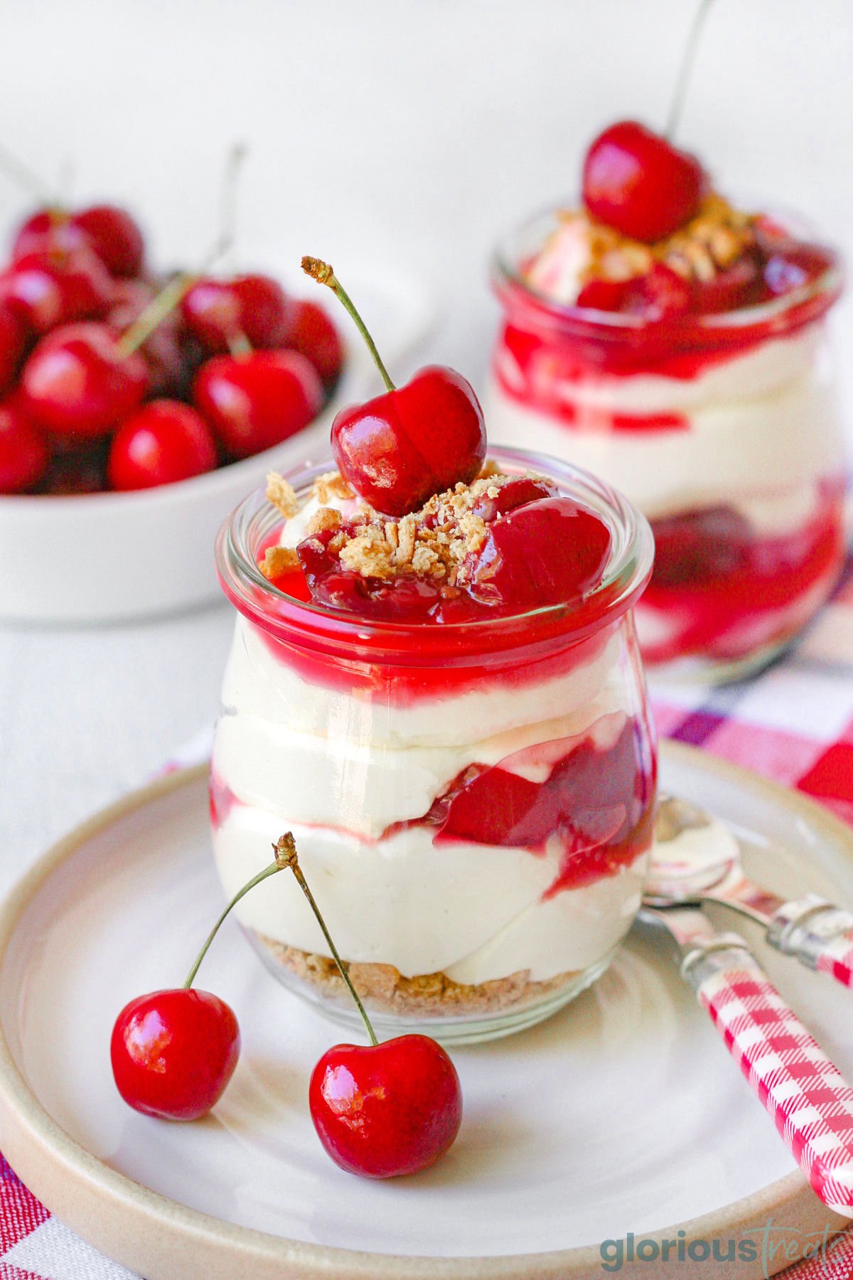 A jar of no bake cherry cheesecake on a white plate with another jar and a bowl full of cherries out of focus in the background.