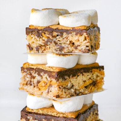 Three decadent smores rice krispie treats stacked on a log. Looking straight on so the layers of rice krispie treats, chocolate, graham crackers, and toasted marshmallows are visible.