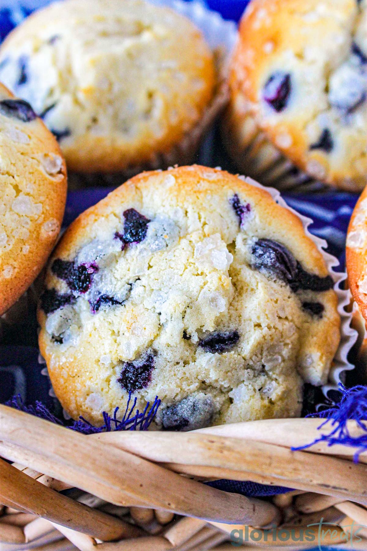 A close up shot of a blueberry muffin in a wicker basket with more muffins behind it.