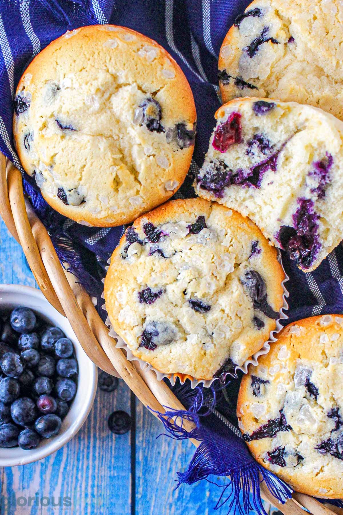Top down look at blueberry muffins in a basket lined with a blue towel. A small bowl of blueberries is off to the side.