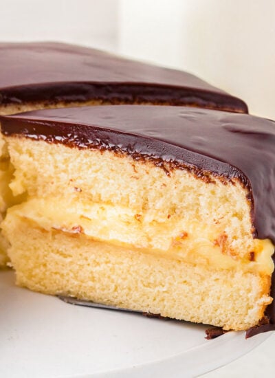 A slice of boston cream pie held on a spatula over a white cake stand.