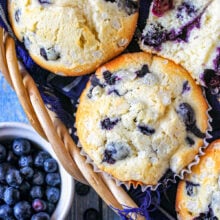Top down look at blueberry muffins in a basket lined with a blue towel. A small bowl of blueberries is off to the side.