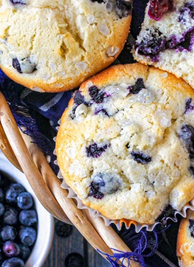 Top down look at blueberry muffins in a basket lined with a blue towel. A small bowl of blueberries is off to the side.
