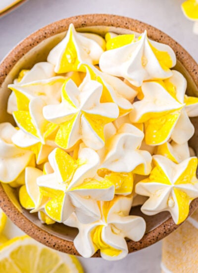 Top down view of lemon meringue cookies in a speckled bowl. Lemons and more cookies are scattered around the bowl.
