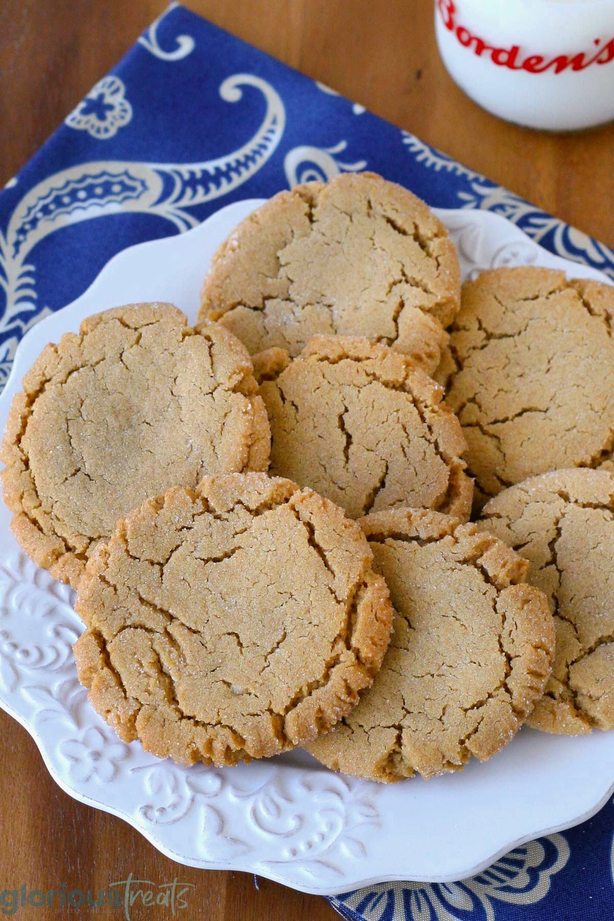 Overhead angled view of white round plate filled with soft and chewy peanut butter cookies next to a jug of milk. Plate is sitting on blue linen napkin.