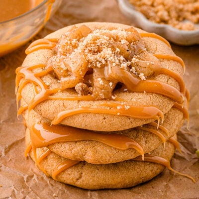 Three caramel apple cookies stacked on brown parchment paper. Caramel drizzle and apple pie filling can be seen on top of the cookies.