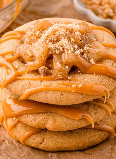 Three caramel apple cookies stacked on brown parchment paper. Caramel drizzle and apple pie filling can be seen on top of the cookies.