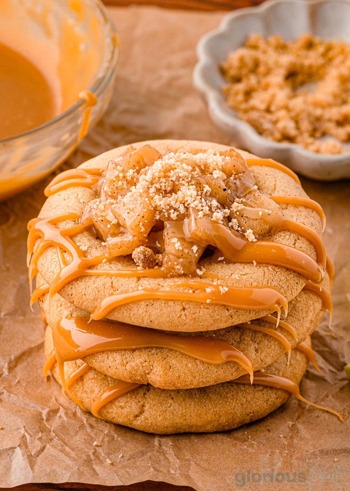 Three caramel apple cookies stacked on brown parchment paper. Caramel drizzle and apple pie filling can be seen on top of the cookies.