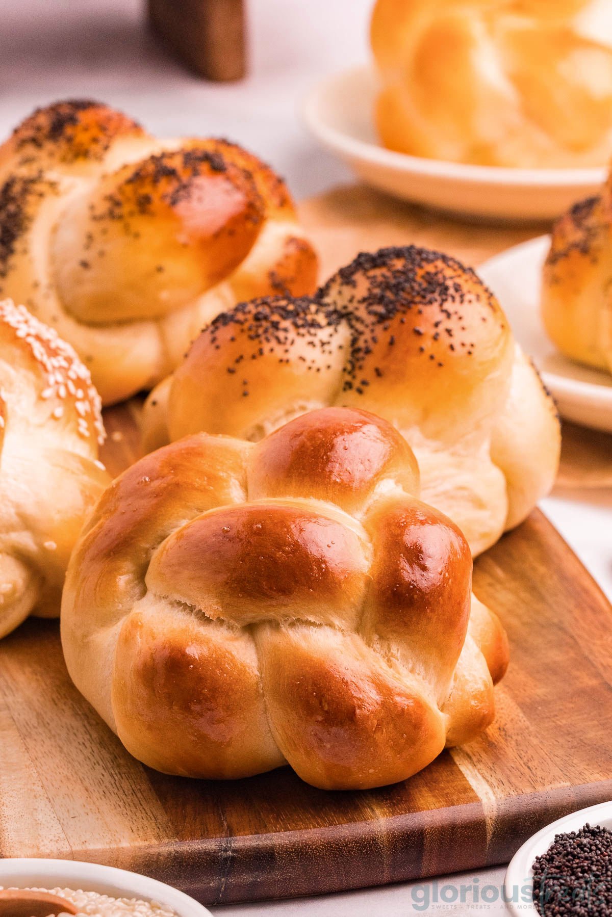 Angled shot of variety of challah rolls on wood board topped with sesame seeds and poppy seeds and plain.