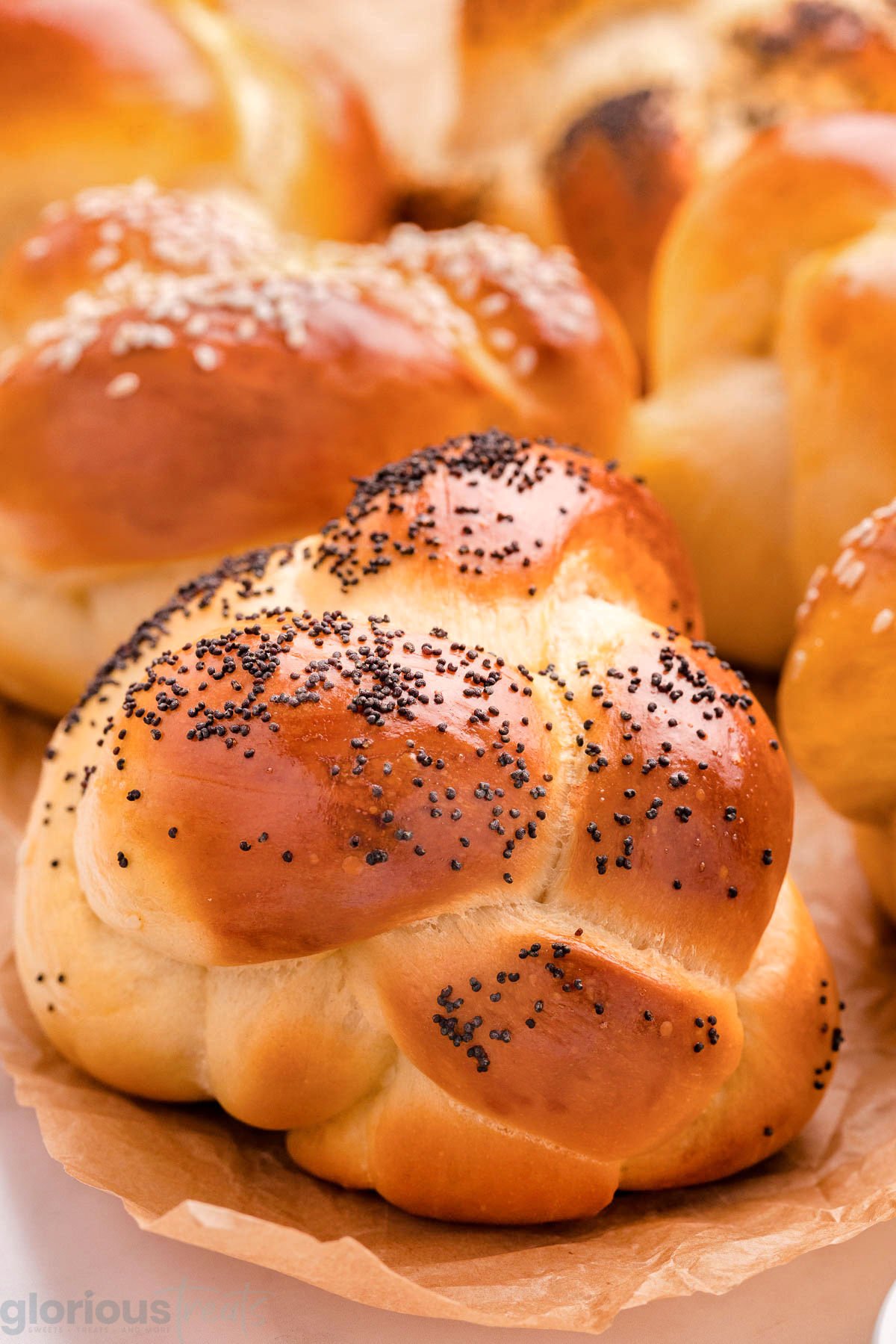 Challah rolls on brown parchment topped with poppy and sesame seeds.
