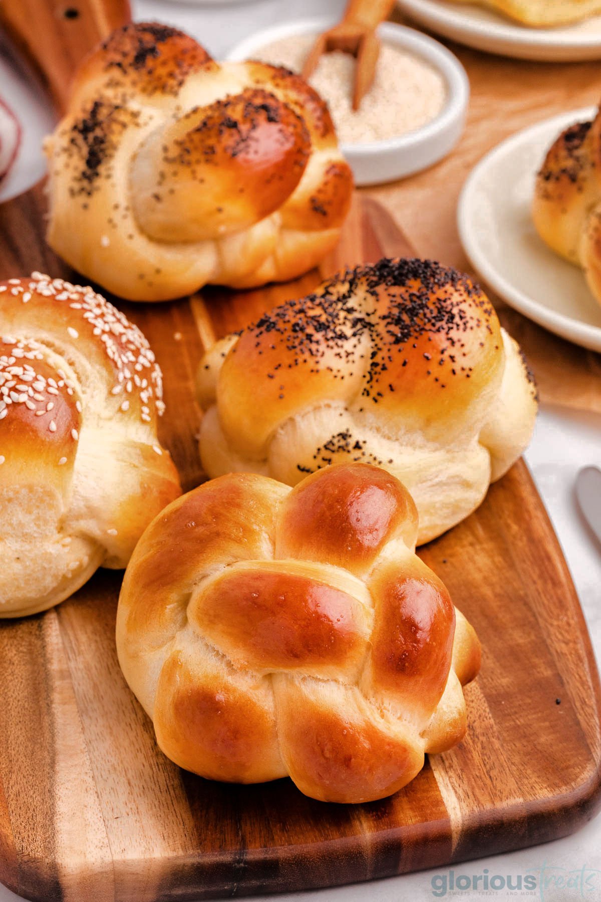 Overhead shot of variety of challah rolls on wood board topped with sesame seeds and poppy seeds and plain.